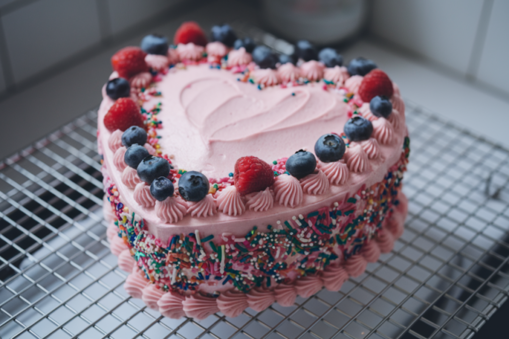 Heart-shaped cake with pink frosting, decorated with sprinkles and fresh berries on a cooling rack.