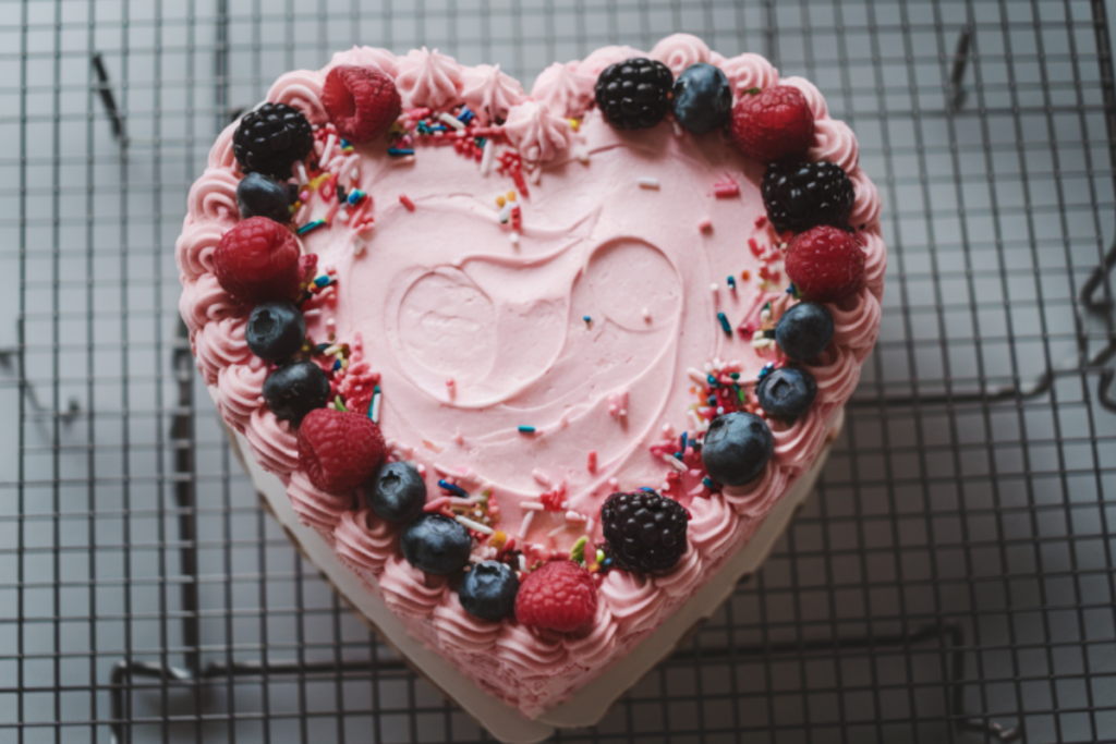 Heart-shaped cake with pink frosting, decorated with sprinkles and fresh berries on a cooling rack.

